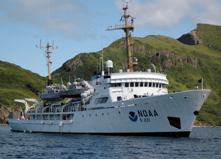 A vessel with a NOAA logo in a body of water with mountains in the background. 