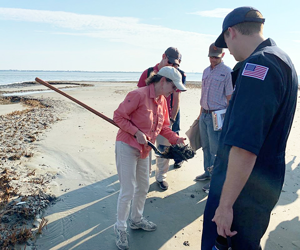 A woman holding a shovel with oiled sand on it for a group of people on a beach to see.