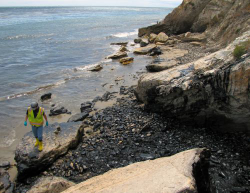 A person walking on a rocky beach with oil on it. 