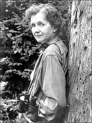 A black and white photo of a woman leaning against a tree.