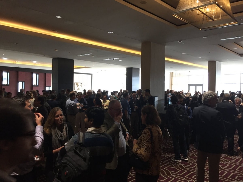 A large group of people standing around talking inside a conference space. 