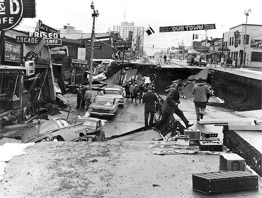 A downtown metropolitan city in 1964 seen with a street collapsed in the aftermath of an earthquake, and community members walking amongst the destruction.