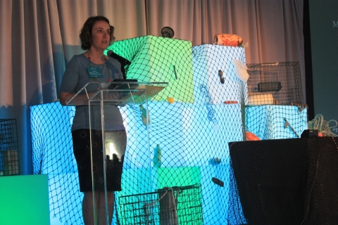A woman at a podium with green and blue light cubes covered in fish netting behind her.