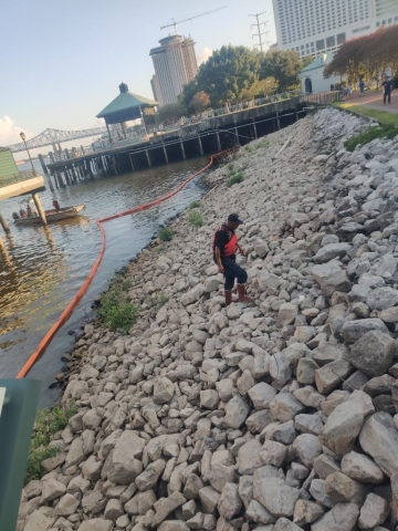 A man in an orange life vest walking up a rocky, urban shore with pollution boom around it.