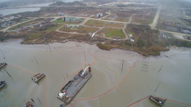 An aerial view of several oil sheens surrounded by pollution boom along a shoreline.