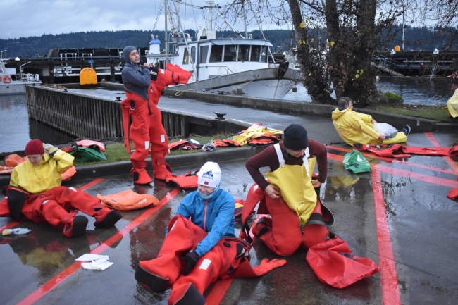 Several people pulling on survival suits on a concrete dock. 