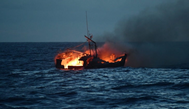 A boat on fire with a smoke plume rising from it. 