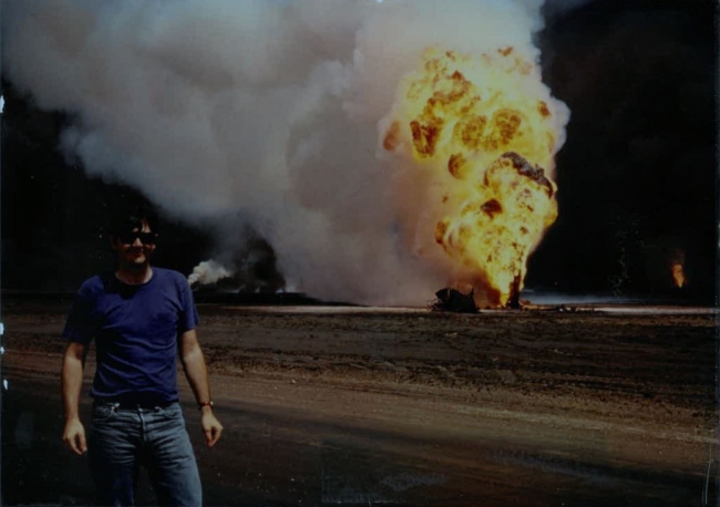 A man posing for a photo with a fire and smoke in the background. 