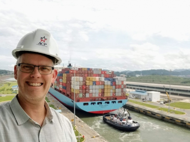 A man wearing a hard hat with a shipping vessel in a lock behind him.