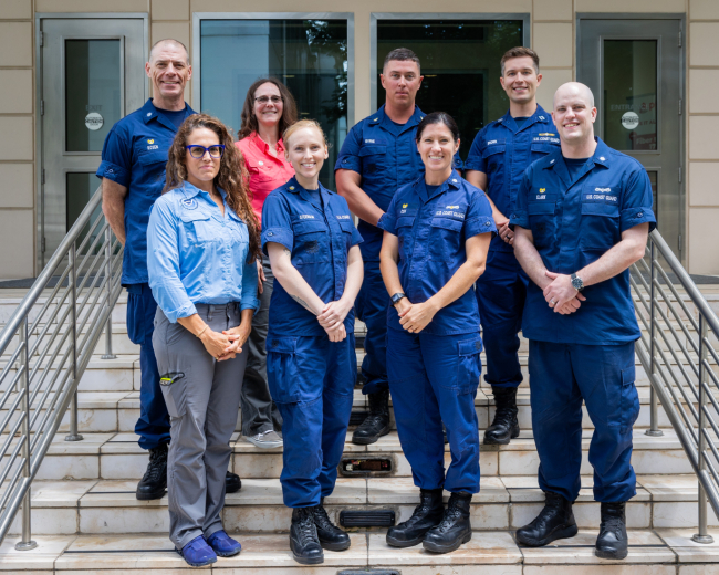 Eight individuals stand together for a group photo on a staircase in front of a building.