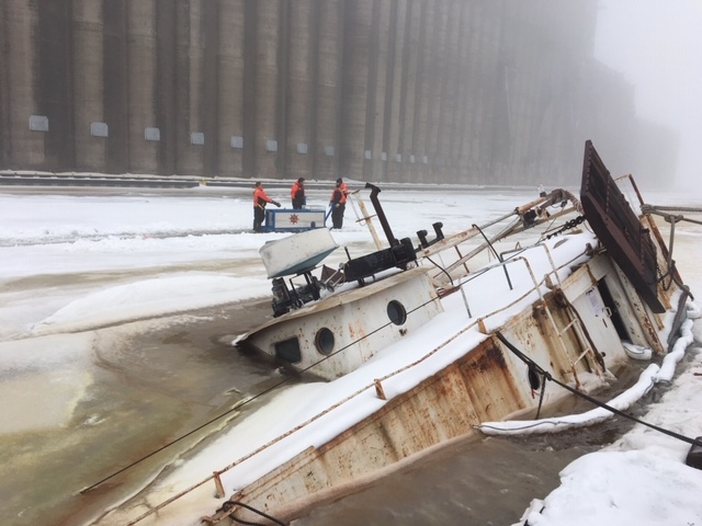 A sunken vessel in an icy-covered body of water with responders in the background. 
