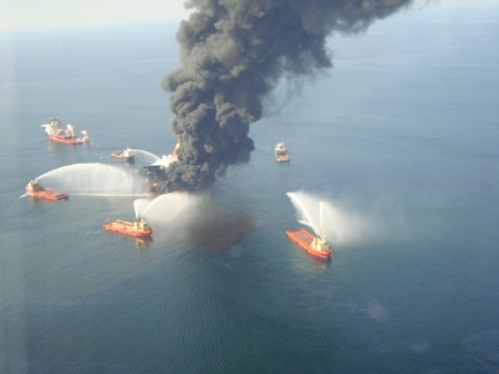 An aerial view of vessels spraying water on a structure with a large plume of black smoke rising from it and visible oil around it. 
