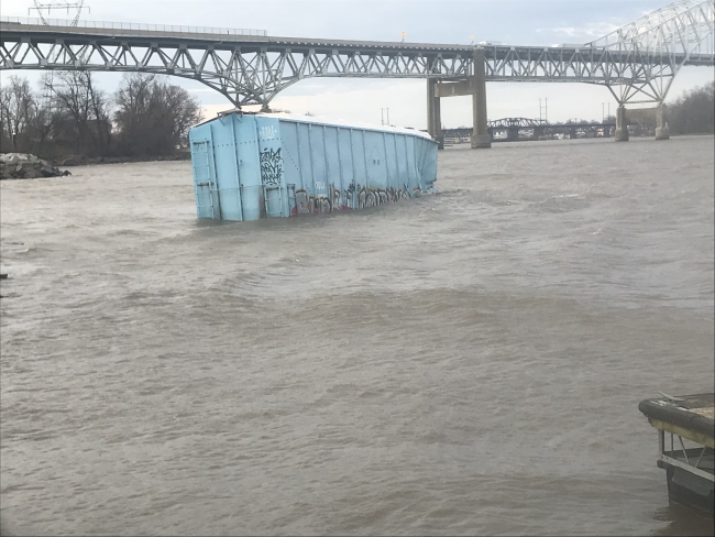 A shipping container in a river with a bridge in the background. 