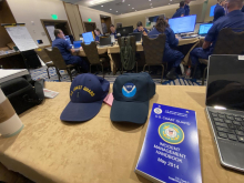 A NOAA hat next to a U.S. Coast Guard hat on a table in a room with responders on their computers.