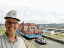 A man wearing a hard hat with a shipping vessel in a lock behind him.