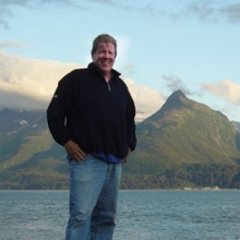 A man posing in front of a body of water and mountains for a photo. 