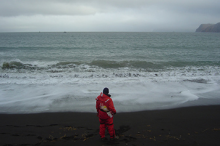 A person in orange gear standing on a beach looking out at the water.