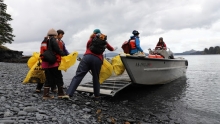 A group of people on a rocky shoreline hauling large, plastic yellow bags onto a boat.