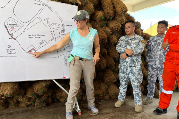 A woman pointing at a map drawn on a white board with men in uniform behind her.